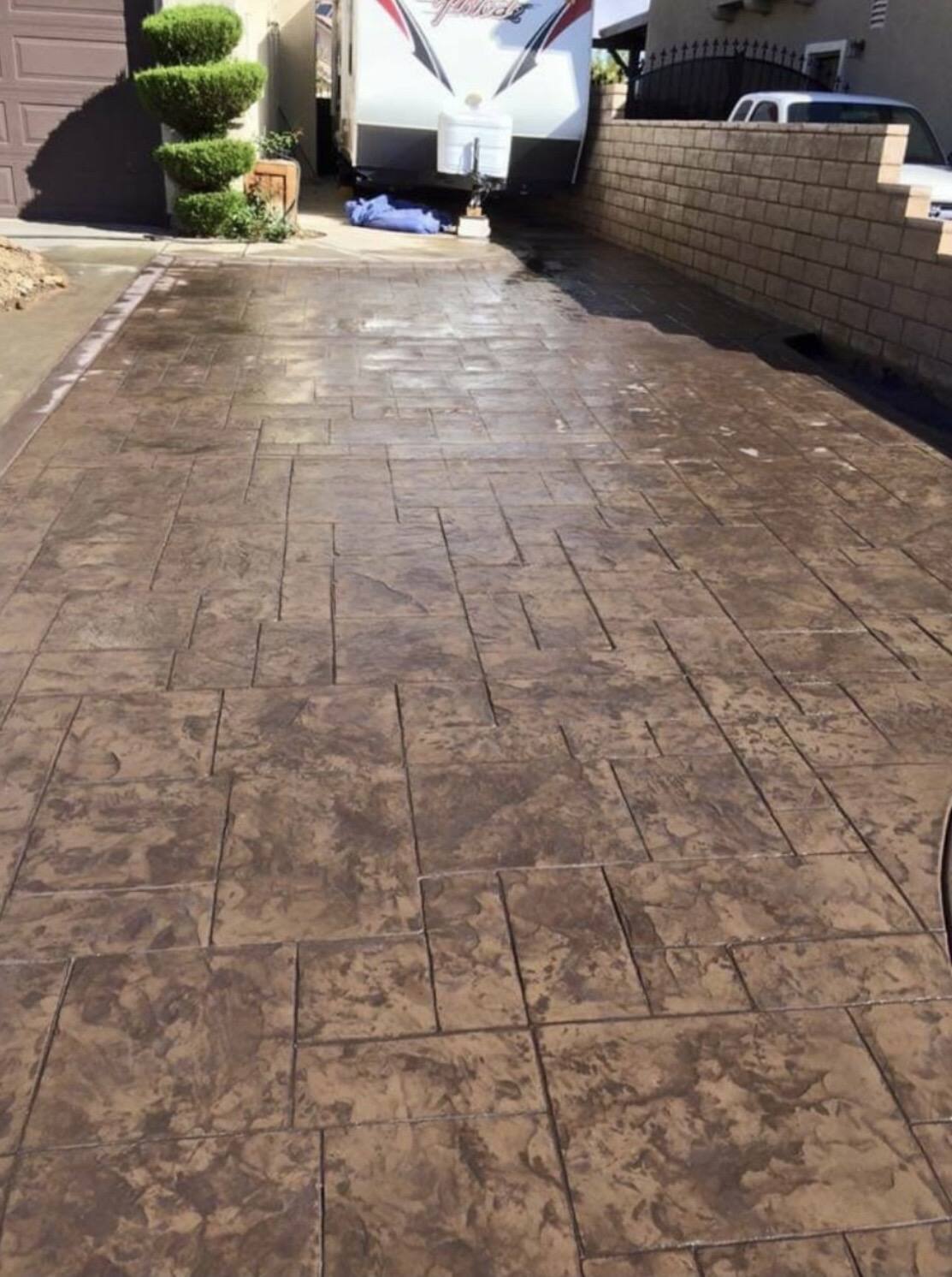 Stamped concrete driveway finished in warm brown with a plank pattern, running between a block wall and a parked trailer