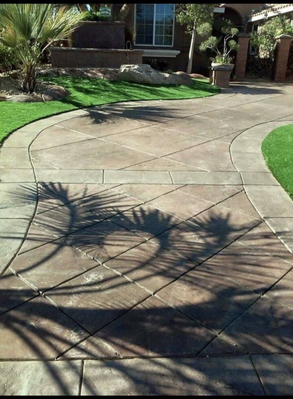 Curved stamped concrete walkway in a tan ashlar pattern, bordered by artificial turf with palm shadows across it